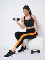 Waylene's high-performance black and mustard gym tights for women, modeled in a gym setting with a woman lifting a dumbbell, focusing on the contrast side stripes and supportive fit.